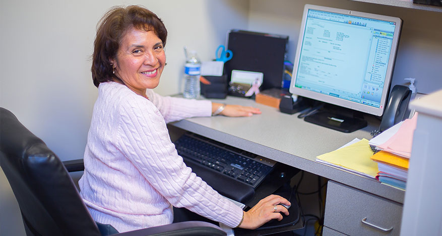 A woman in a light pink sweater sits at a desk, smiling at the camera, while working on a desktop computer. The desk has papers, folders, a water bottle, and office supplies.