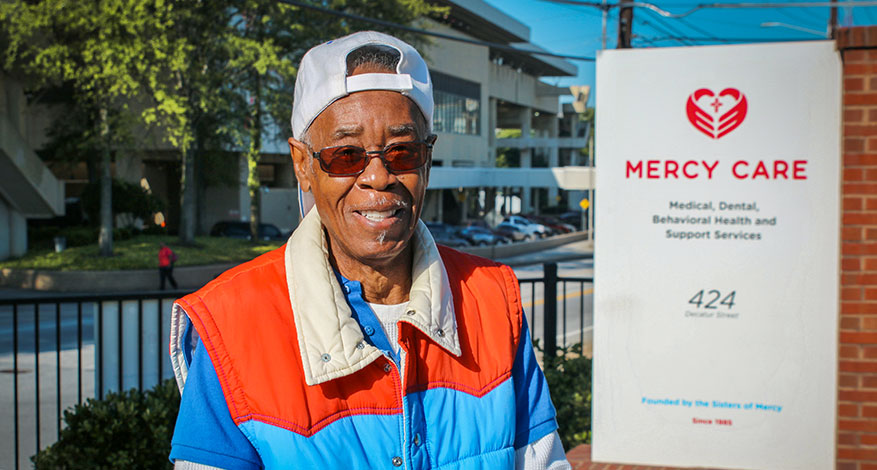 An older man with swagger, wearing a white cap, sunglasses, and a colorful vest, smiles while standing outside next to a sign for Mercy Care, a medical and support services facility.
