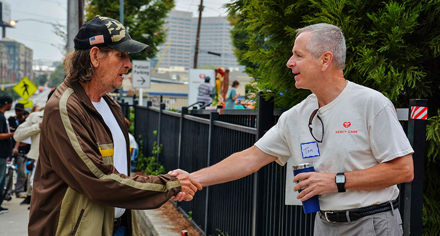 Two men shake hands outdoors near a black metal fence; one wears a cap and casual jacket, the other wears a “Mercy Care” shirt and holds a blue cup. People and buildings are visible in the background.