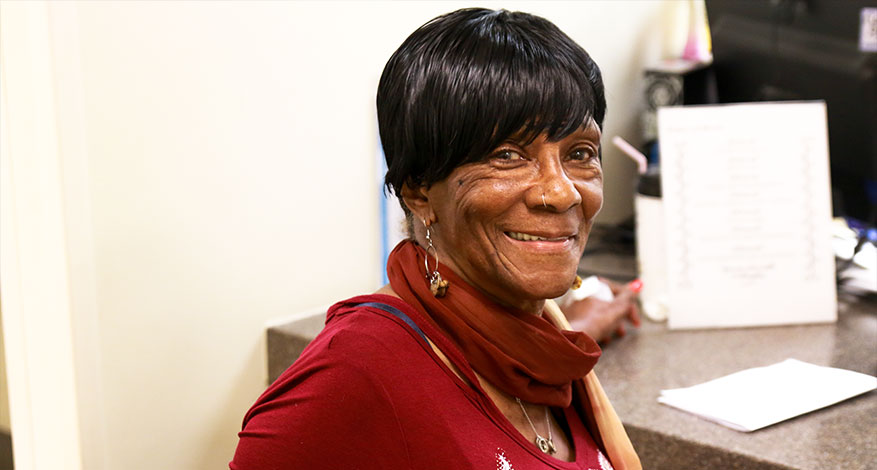 An older woman with short black hair, wearing a red top and scarf, smiles warmly while standing near a countertop in an office setting, showing her love for helping people every day.