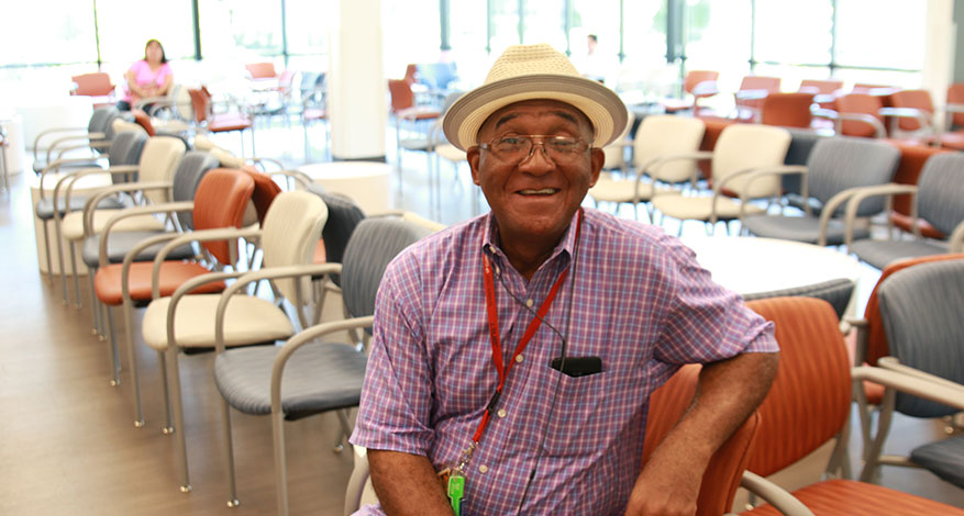 An older man wearing a straw hat, glasses, and a purple plaid shirt shares a meaningful smile while sitting in a bright, spacious waiting room filled with colorful chairs.