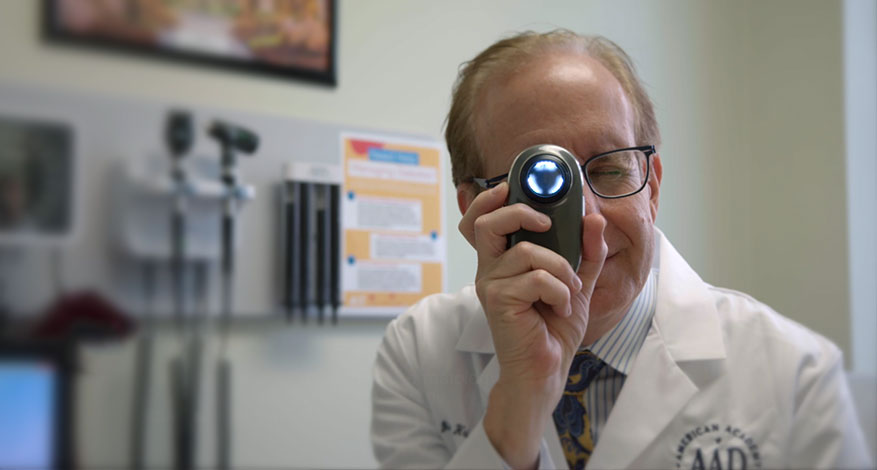 A doctor in a white coat examines a patient using a handheld medical device with a bright light in a dermatology clinic, surrounded by medical equipment in the background.