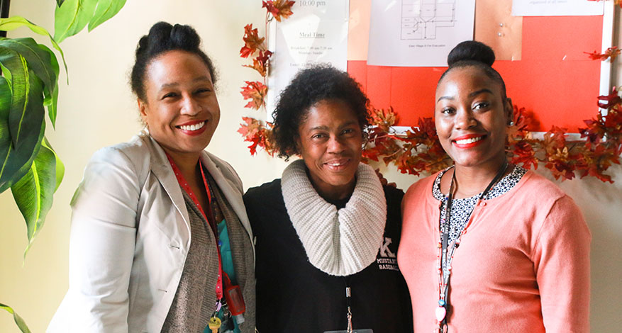 Three women stand close together, smiling just for me at the camera in an indoor setting decorated with autumn leaves and a bulletin board in the background.
