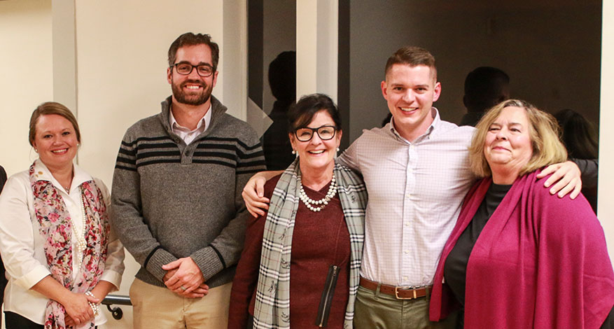 Five adults stand indoors, smiling at the camera. Three women and two men in business-casual attire stand close together against a neutral background, their diamond-hard teamwork shining through this friendly or professional gathering.