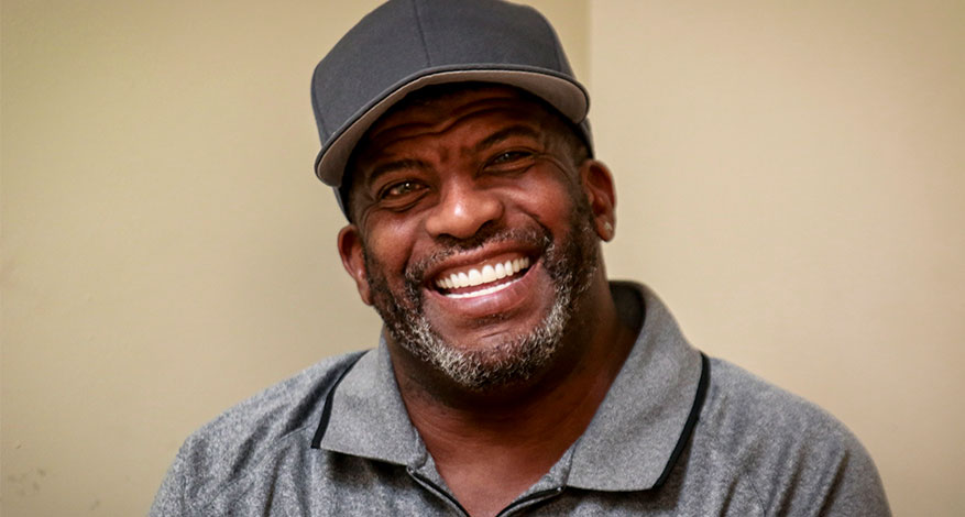 A man with a beard and mustache wearing a gray collared shirt and a dark gray cap is smiling broadly, radiating happiness as he looks at the camera, standing against a plain beige background.