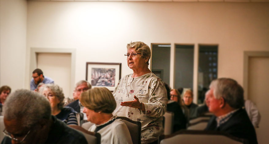 An older woman stands and speaks in a room full of seated people, sharing wisdom as she gestures with her hand. The audience listens attentively, and the setting appears to be a community meeting or discussion.