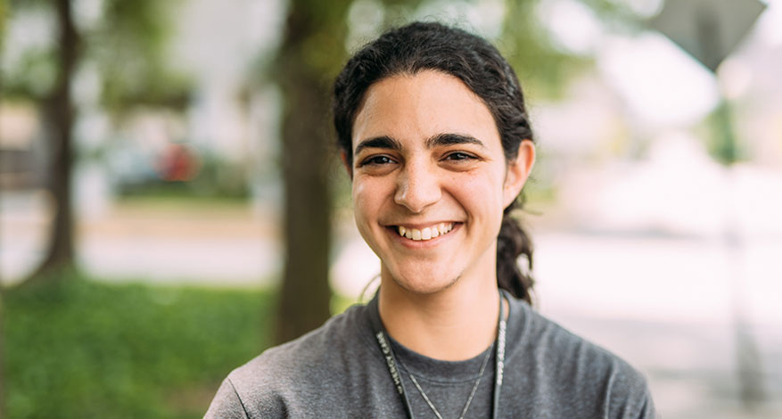 A young person with long dark hair tied back, wearing a gray t-shirt and a necklace, smiles outdoors with greenery and trees softly blurred in the background, inspired to help keep people alive through kindness and compassion.