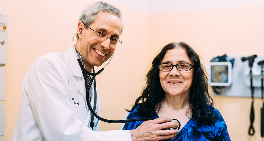 Dr. Onofre smiles while using a stethoscope to listen to a female patient’s chest. The patient is also smiling as they sit together in a medical examination room with equipment visible in the background.