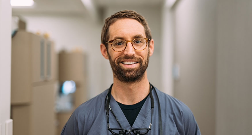 A smiling man with glasses and a beard, wearing scrubs and a stethoscope, stands in a well-lit hallway, embodying the aspirations of what I want to be in a medical or healthcare facility.