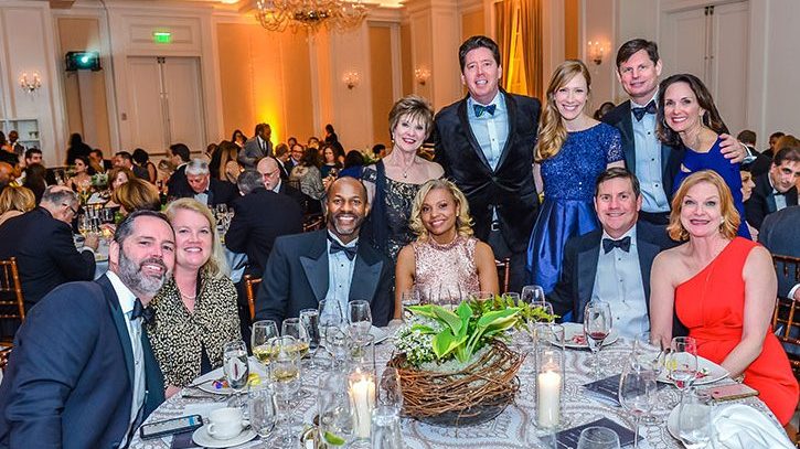 A group of elegantly dressed people, posing and smiling around a round table at the Mercy Ball 30th Anniversary, with dinnerware, glasses, and a floral centerpiece in a bright, decorated ballroom filled with other guests.