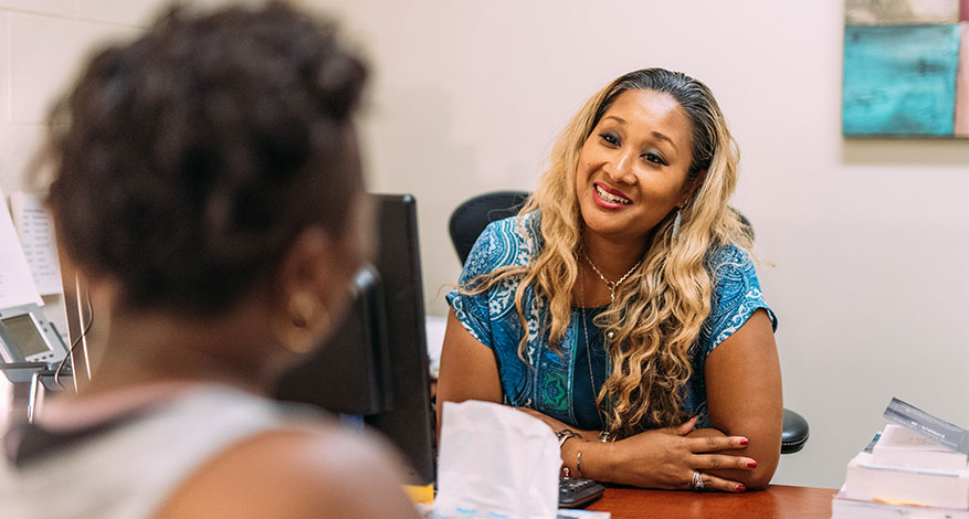 A smiling woman with long wavy hair, wearing a blue top, sits at a desk and discusses overcoming adversity with another person whose back is to the camera in an office setting.