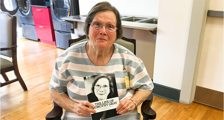 An elderly woman wearing glasses and a striped shirt sits in a chair, smiling and holding a book titled The Life of Dorothy Gray, which features her photo on the cover—a cherished family heirloom in an inviting indoor common area.