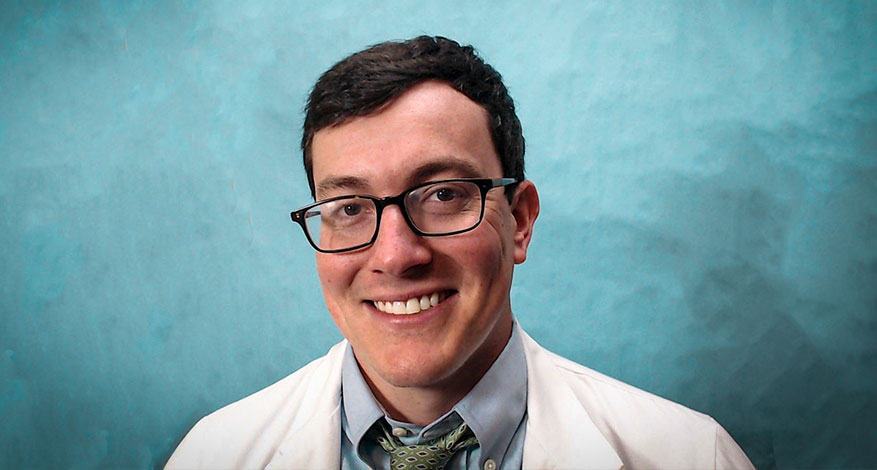A man with short dark hair and glasses smiles at the camera. He is wearing a white lab coat, a blue dress shirt, and a patterned green tie, standing in front of a teal background.