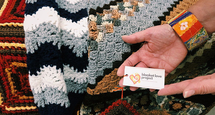 A hand wearing a colorful embroidered bracelet holds a “Blanket Love Project” card next to several crocheted blankets, showcasing geometric patterns and vibrant colors that celebrate tradition and warmth.