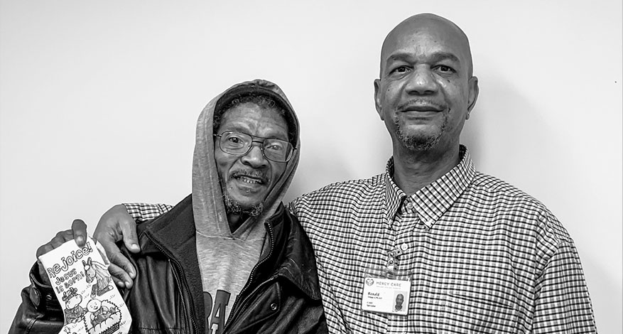 Two men stand together smiling in front of a plain wall. The man on the left wears glasses, a hoodie, and a leather jacket, holding a card as if showing step by step progress. The man on the right wears a checked shirt and an ID badge.