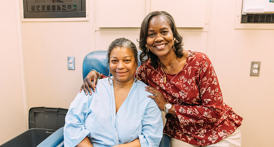 Two women smiling in a medical setting; one is sitting in a hospital gown while the other stands beside her with an arm around her shoulders, both looking at the camera—capturing a joyful moment from Tammy's story.