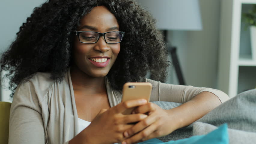 A woman with curly hair and glasses is smiling while looking at her smartphone, enjoying Behavioral Health visits from home as she sits on a couch in a cozy indoor setting.