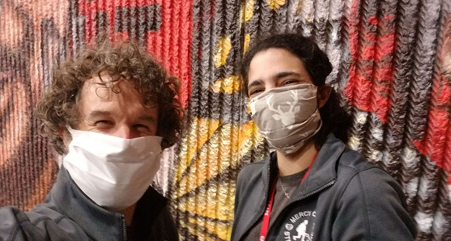Two people wearing face masks and dark jackets, part of local outreach teams, stand in front of a vibrant mural. Both are smiling under their masks, reflecting positivity during the COVID-19 response. The mural features bold red, yellow, and black hues.