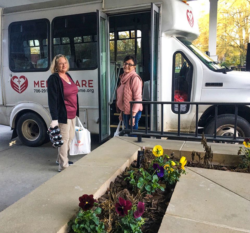 Two women stand outside a white van with a Medicare logo, finding their way amid colorful flowers in the foreground. One holds water bottles and a bag, while the other waits by the van door, as if ready for a journey to Rome.
