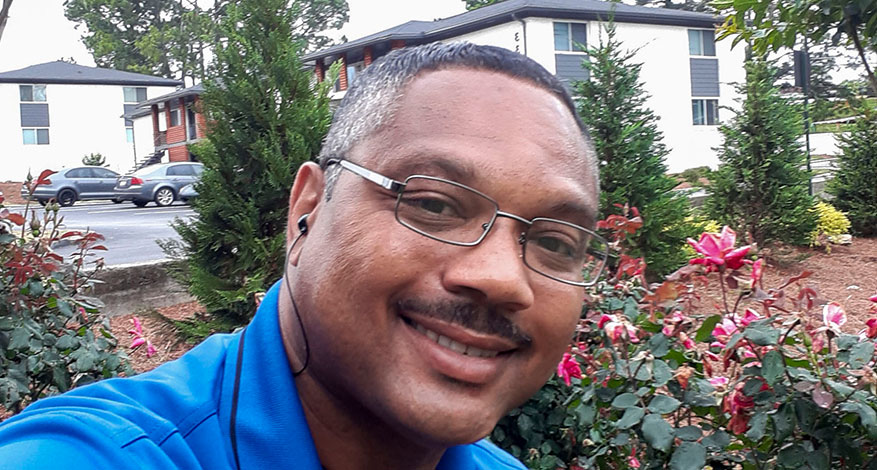 A man wearing glasses and a blue shirt smiles at the camera while sitting outdoors near blooming pink flowers, with a straight path, apartment buildings, and parked cars in the background.