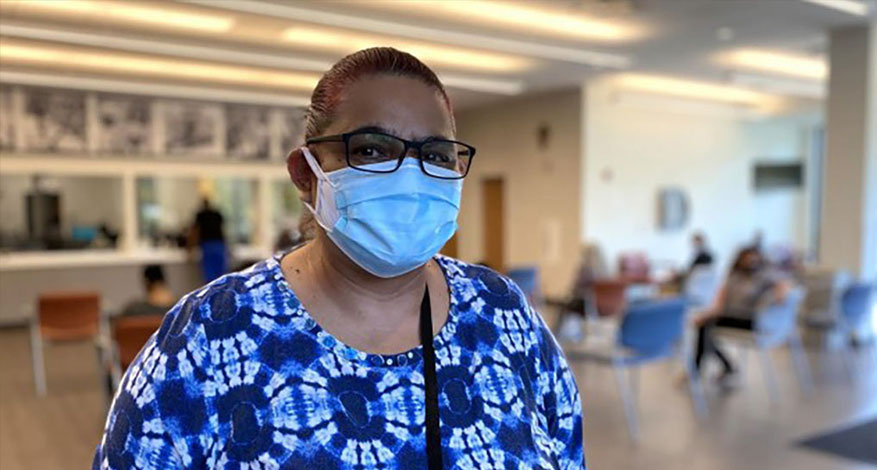 Hortensia, wearing glasses, a blue and white patterned top, and a face mask, stands in a well-lit waiting area—an important step in her health journey—while several seated people are blurred in the background.