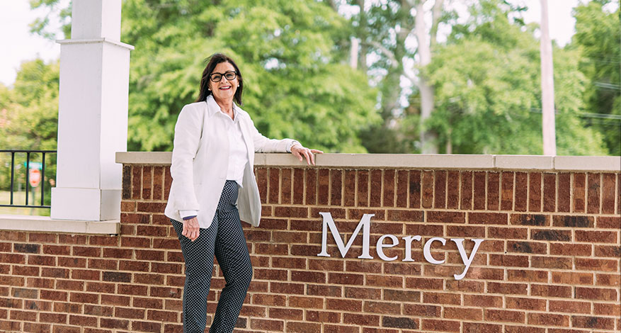A woman in a white jacket and patterned pants stands and smiles by a brick wall with the word Mercy on it, reflecting the community impact of Mercy Care in Rome. Trees and greenery can be seen in the background.