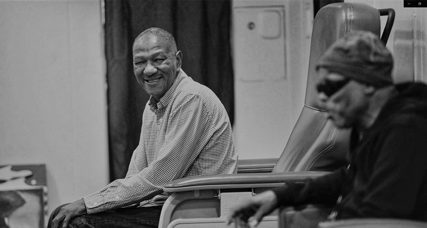 Two men sit indoors; one, in a checkered shirt, smiles and looks toward the other, who wears a hooded jacket and sunglasses, seated in a reclining chair. The black-and-white scene quietly reflects the spirit of recuperative care.