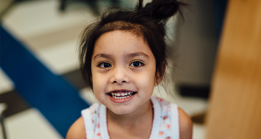 Young girl with dark hair in a top knot smiles brightly at the camera, wearing a white floral top. The background is softly blurred in blue and beige tones, creating a warm Hearts and Hands atmosphere.