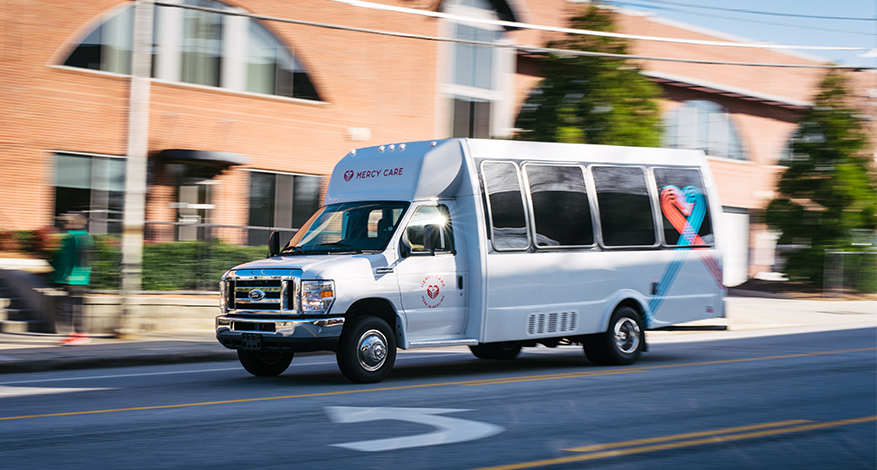 A white passenger shuttle bus with Vekzy Care branding drives on a city street, with blurred buildings and people in the background, suggesting motion—a scene reminiscent of Ruben's Story.