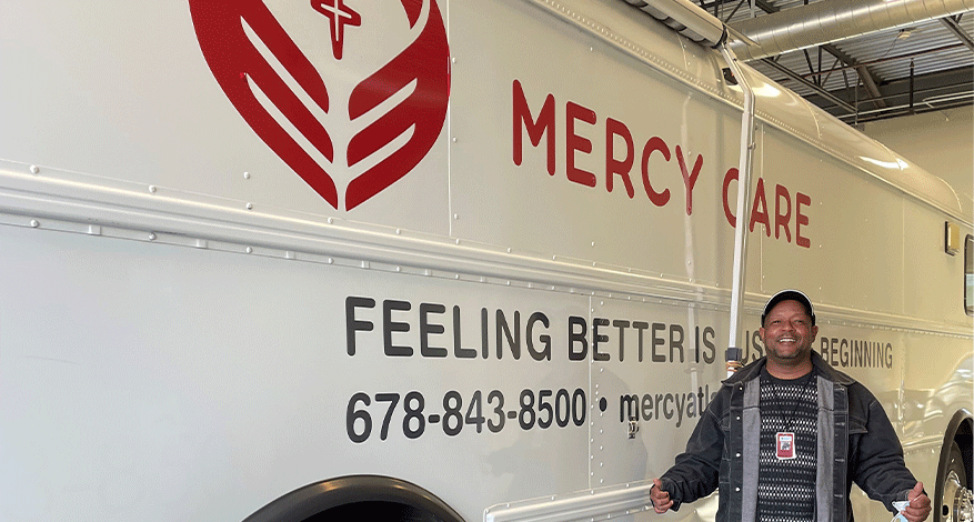 A smiling man stands in front of a white Mercy Care mobile clinic van with a large red heart logo, text reading “FEELING BETTER IS JUST THE BEGINNING,” and a phone number on the side.