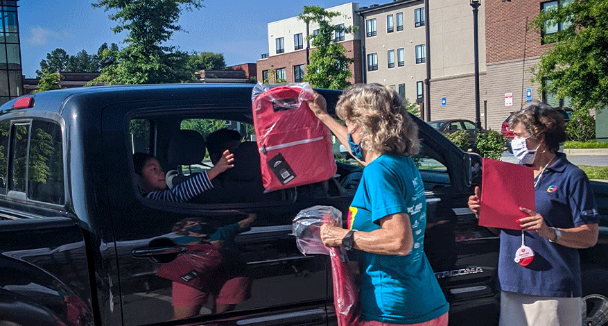 Two women hand out a versatile backpack to a child in a black pickup truck during a daytime outdoor event, with apartment buildings and trees visible in the background.
