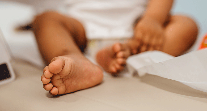 A close-up of a young children's bare feet as they sit on a light-colored surface, with their legs crossed and a hand resting on one knee. The focus is on the feet, while the background is softly blurred.