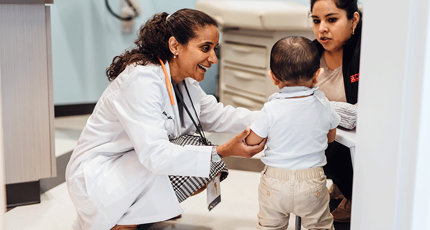 A doctor kneels and smiles while examining a toddler who stands facing her, with the child’s mother sitting nearby in the clinic room—a warm example of families connecting in their trusted medical home.