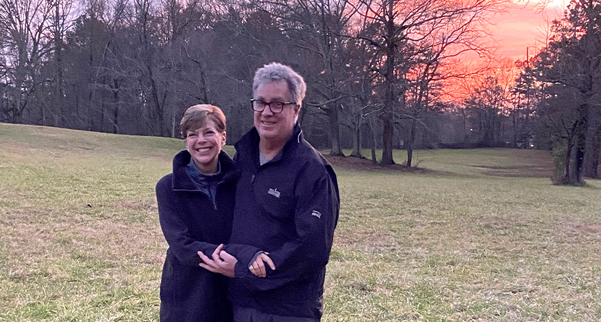 A smiling couple stands close together in an open grassy field at sunset, the colorful sky behind them—a perfect moment that feels like a gift that keeps on giving. Both wear dark jackets and glasses, framed by leafless trees in the background.