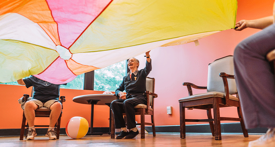 Several older adults at Mercy Care Rome sit indoors, joyfully lifting a colorful parachute together, with a beach ball and sunlight streaming in—celebrating hope and connection in healthcare.