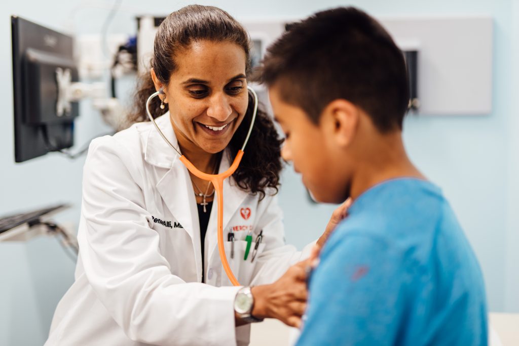 A female doctor in a white coat uses a stethoscope to examine a young boys chest in a medical office, both smiling and engaged in the check-up.
