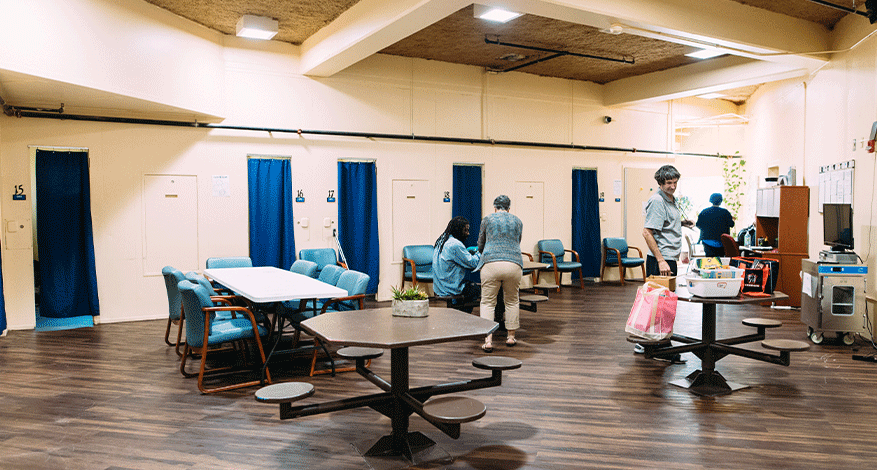 A clinic waiting area with several empty chairs, tables, and blue privacy curtains. A few people are standing and talking, building long lasting friendship as medical staff attend to equipment carts. The room has wood flooring and cream-colored walls.