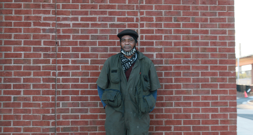 A man wearing a green jacket, scarf, and hat stands against a red brick wall with his hands in his pockets, looking at the camera—capturing the relief that followed his recent life saving diagnosis.