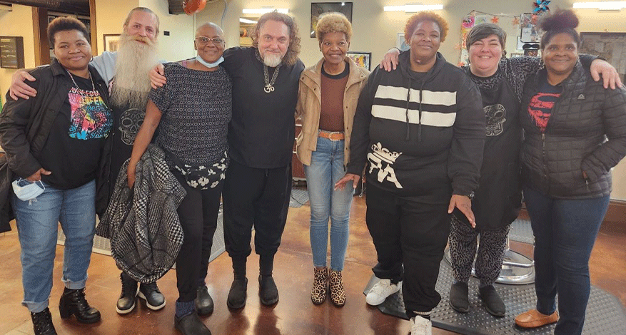 A group of eight people stand closely together indoors, smiling at the camera. Dressed casually, they appear to be in a friendly, relaxed setting with salon chairs and decor at Decatur Barbers, where kindness and great haircuts go hand in hand.