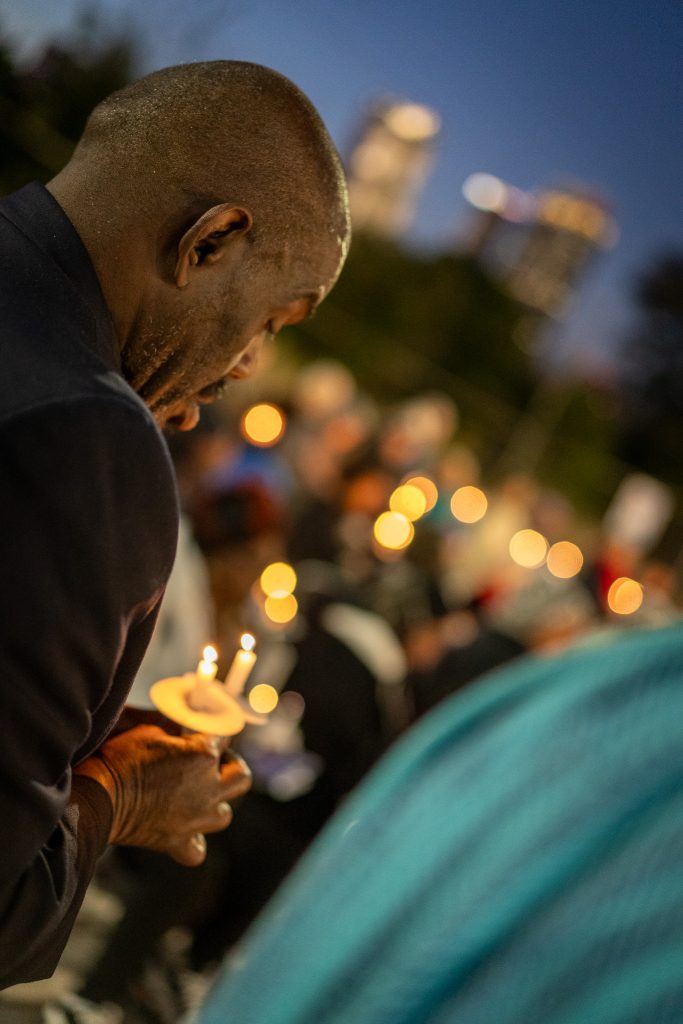 A man holds a lit candle during the Annual Memorial vigil at dusk, surrounded by others honoring loved ones. The background glows softly with warm lights, city buildings, and the spirit of Mercy Care.