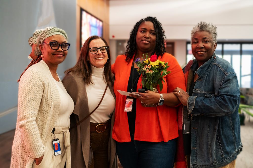 Four women smiling and standing together indoors, one holding a bouquet of flowers and an envelope, celebrate what appears to be a special event—possibly the McAuley Award at Mercy Care—with Beverly Kerr-Caesar among them.