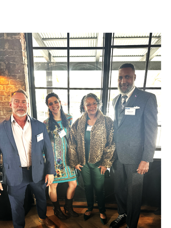 Four adults, three in suits and one in a patterned dress, stand together indoors by large windows. The person second from the right wears a leopard print jacket. All are smiling—true Healthcare Champions from the Mercy Care Street Medicine Team.