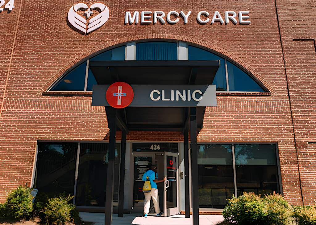 A person enters a brick building with signs reading MERCY CARE and CLINIC above the entrance. The clinic has a red medical cross symbol and large windows reflecting the outside.