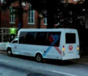 A white shuttle bus with blue and red heart-shaped graphics on its side and back is parked on a city street near a brick building.