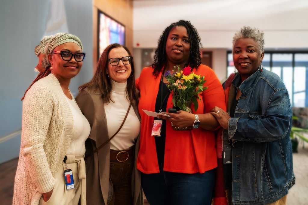 Four women stand together indoors, smiling at the camera. One woman holds a bouquet of flowers and an envelope. They appear to be in a celebratory or professional setting, with large windows and screens in the background.