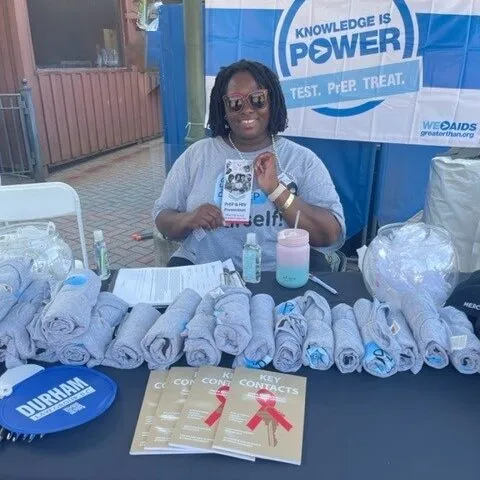 A woman sits at a table promoting HIV awareness, holding a pamphlet about PrEP. The table is covered with towels, flyers, and informational materials. A banner behind her reads Knowledge is Power: Test. PrEP. Treat.