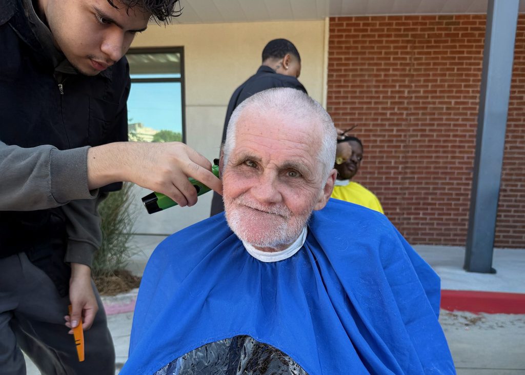 An older man with short gray hair and a beard smiles while getting a haircut outdoors from community volunteers. He wears a blue barber cape as his hair is trimmed. Two people are seen in the background near a brick building.