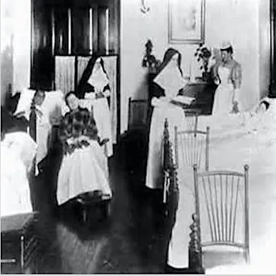 Black and white photo of nurses and nuns caring for patients in beds in a hospital room, likely from the early 1900s. The room has wooden floors, chairs, and partly screened-off beds.