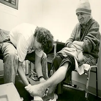 A person sits on a chair with a blanket over their lap while another person kneels, tending to their bare feet. The setting appears to be a simple room, and the atmosphere suggests care and assistance.