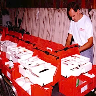 A man in a white shirt stands next to several open red toolboxes or medical supply cases, sorting or organizing various items inside them. The background shows beige curtains.